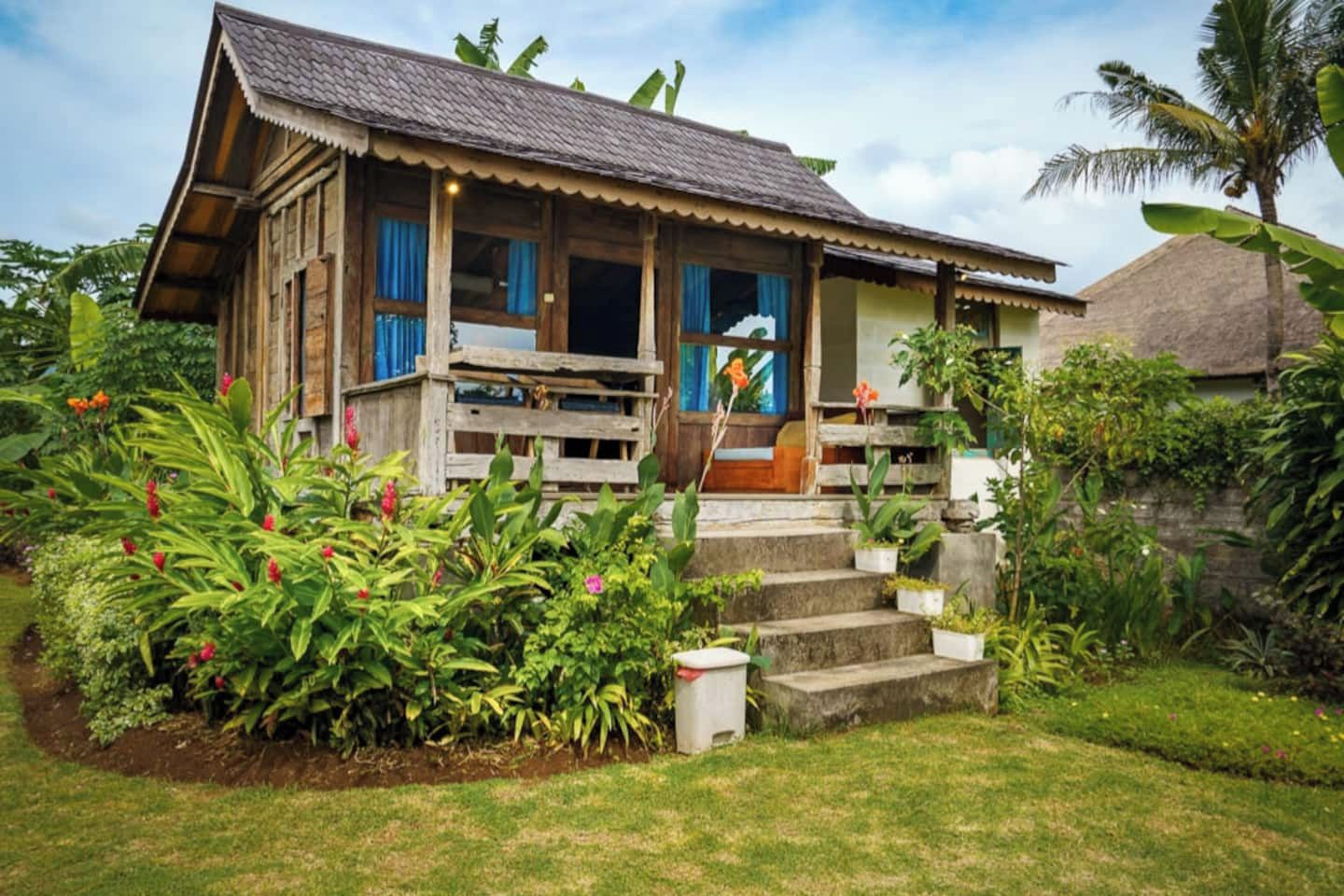 A wooden house with a porch, surrounded by lush greenery and a small garden.