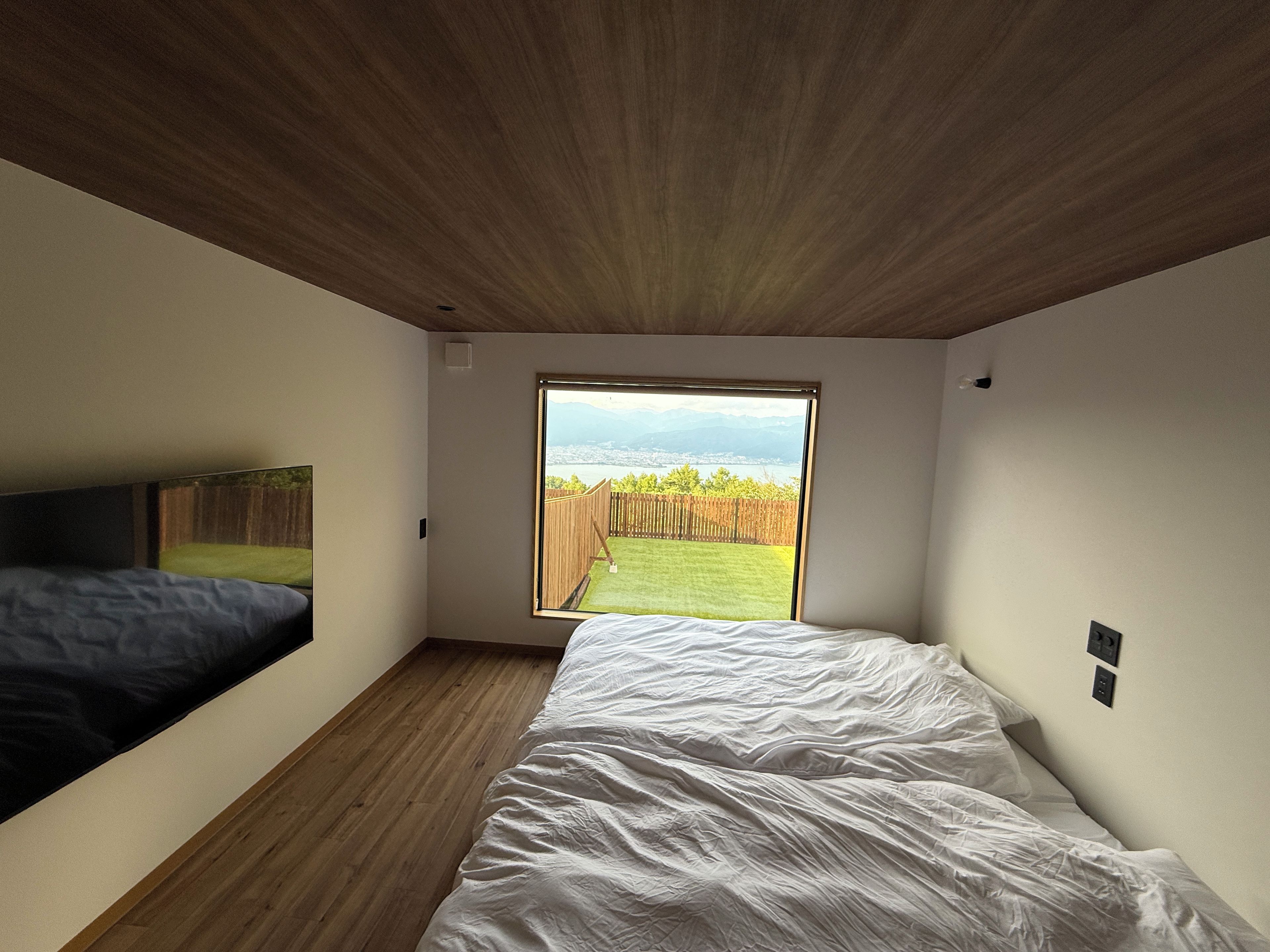 A bedroom with a large window, a bed, and a wooden ceiling.