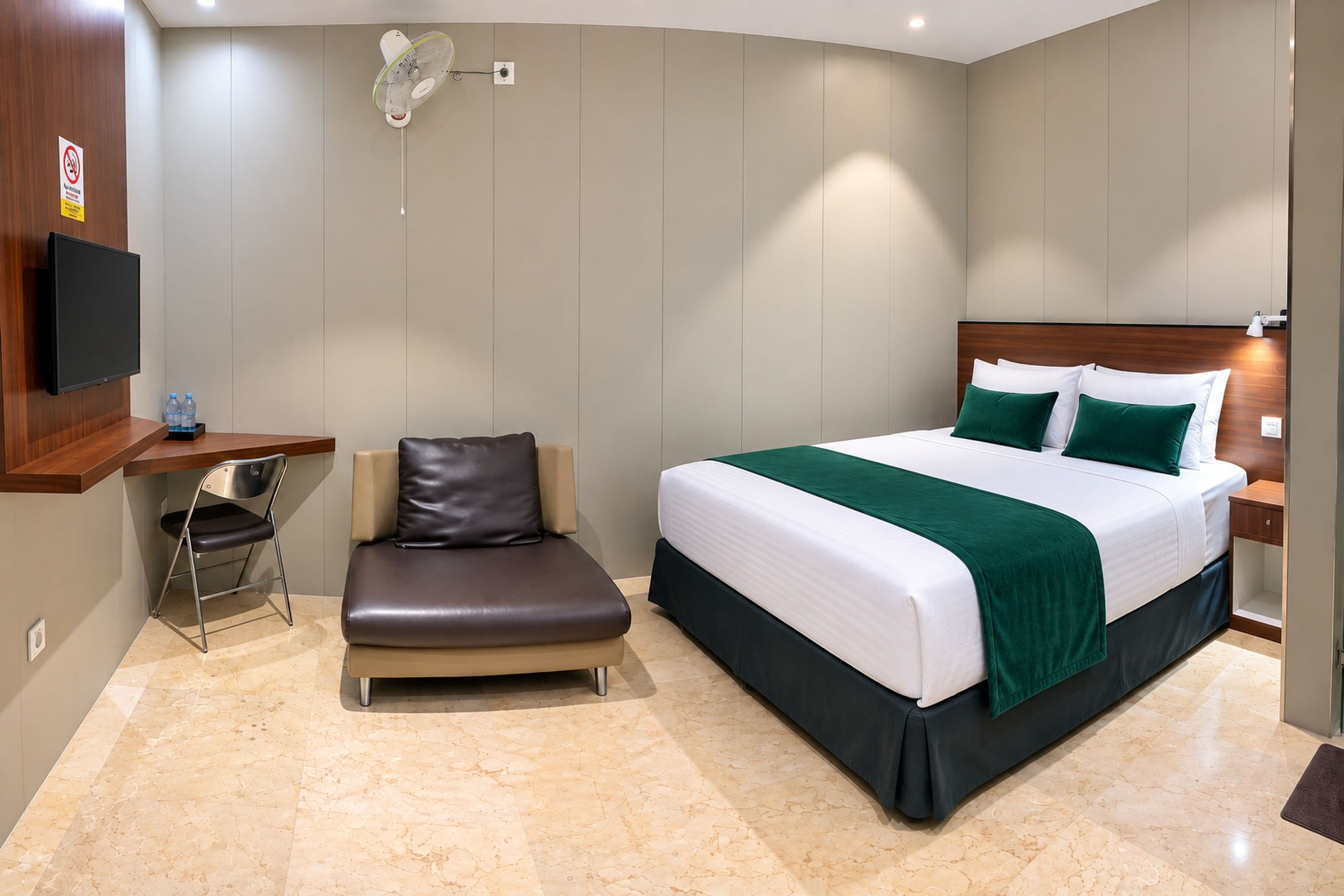A hotel room with a bed, a desk with a chair, a standing fan, and a television.