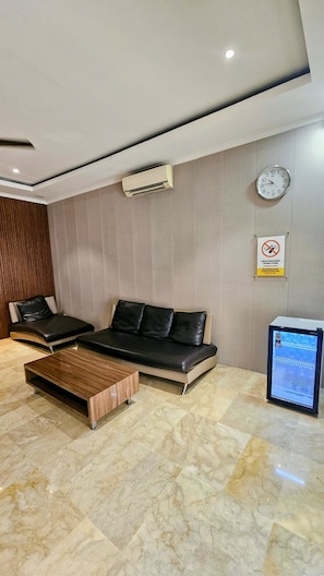 A modern waiting area with a sofa, a wooden coffee table, a wall-mounted clock, an air conditioning unit, and a digital screen displaying a map. - 555 Nangka Guest House (Jakarta Selatan)