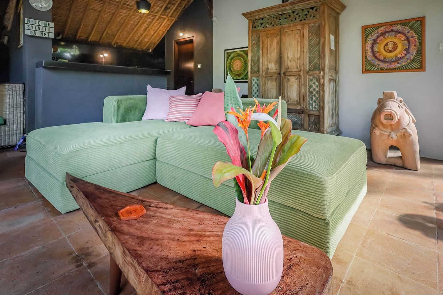 A green sofa with pillows, a wooden coffee table, a vase with flowers, a wooden cabinet, and a decorative piggy bank.