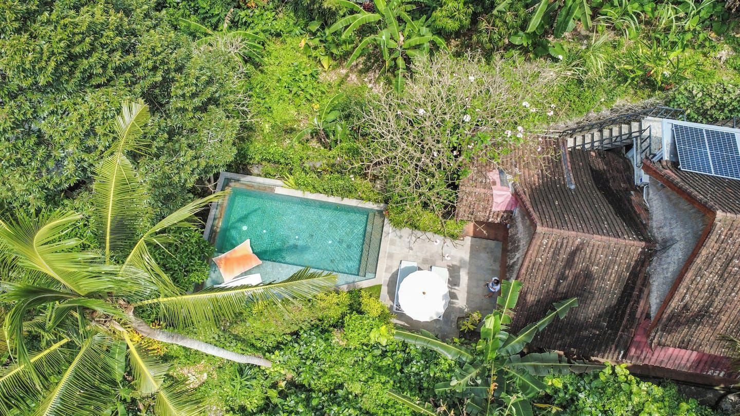 An aerial view of a property with a swimming pool surrounded by lush greenery, a covered patio area, and a solar water heater.