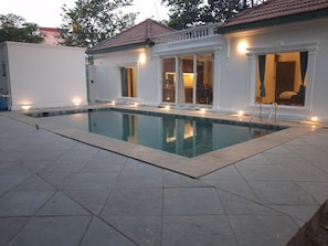A swimming pool with a tiled deck, a white building with a red roof, and exterior lighting.