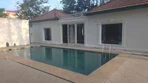A rectangular swimming pool with a tiled deck, surrounded by a white building with a red-tiled roof and large windows. - The Cosy Pool Villa by Azure Stays (Mahabalipuram)