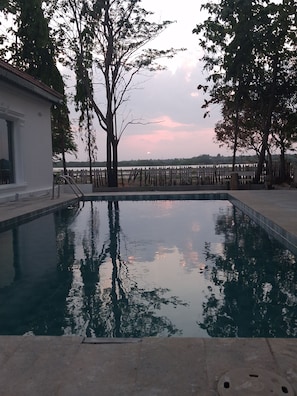 A swimming pool with a wooden fence, trees, and a view of the water body in the background. - The Cosy Pool Villa by Azure Stays (Mahabalipuram)