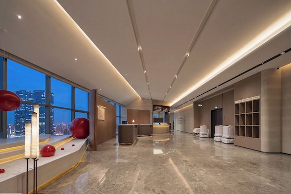 Un moderno vestíbulo de hotel con mostrador de recepción, zona de entrega de comida y vistas a la ciudad a través de amplias ventanas.
