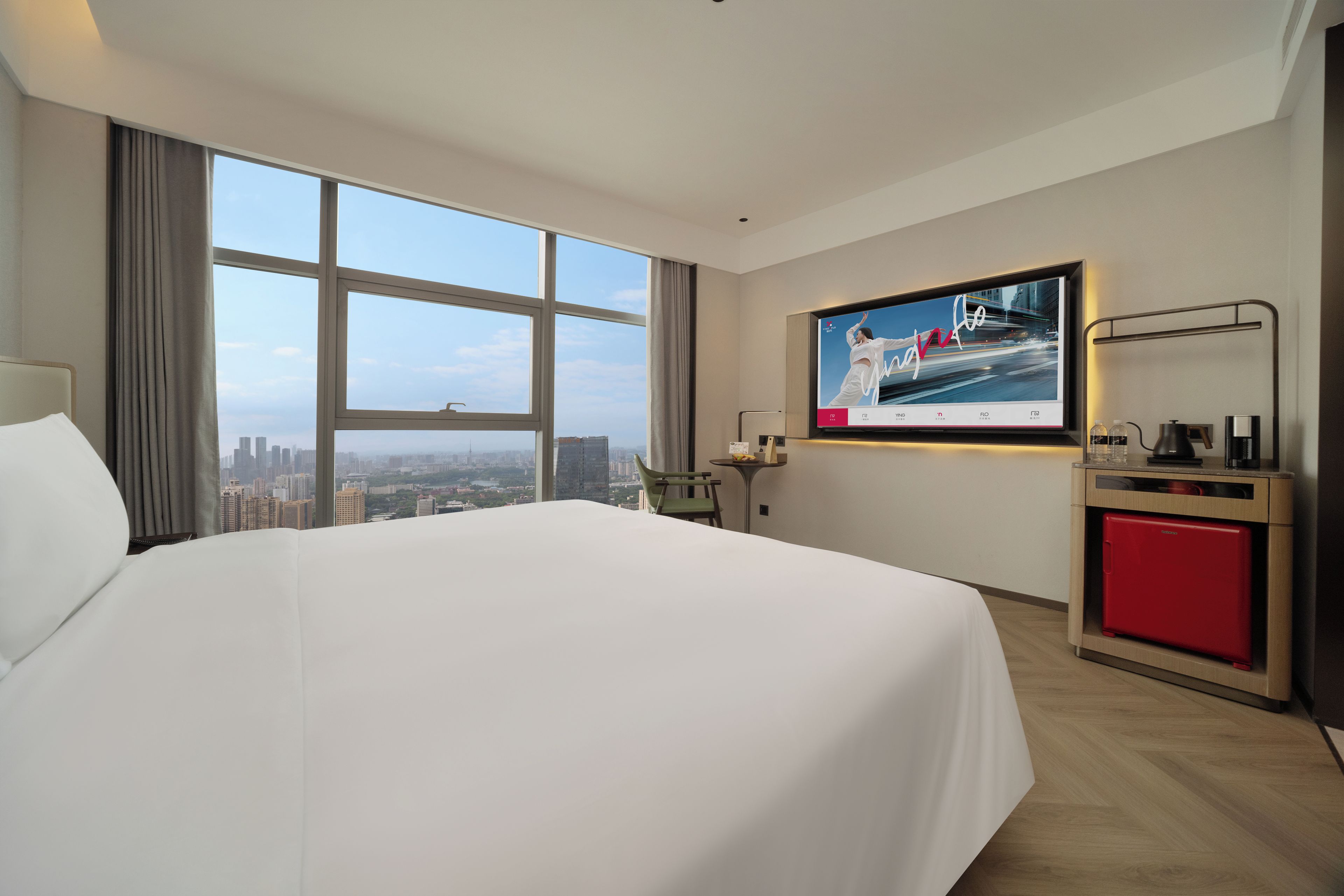 A hotel room with a large bed, a TV mounted on the wall, a small table with a chair, and a view of the cityscape through large windows.