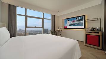 A hotel room with a large bed, a TV mounted on the wall, a small table with a chair, and a view of the cityscape through large windows.