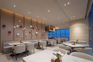 Interior moderno de un restaurante con múltiples mesas y sillas, vistas a la ciudad a través de grandes ventanales y luces esféricas decorativas en la pared.
