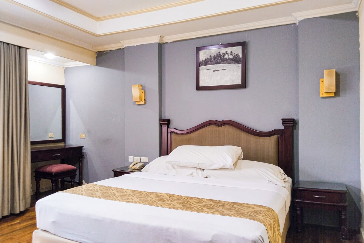A hotel room with a large bed, a bedside table, a desk with a chair, a mirror, and a framed picture on the wall.