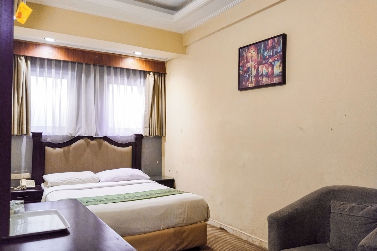 Habitación de hotel con cama, escritorio, silla, un cuadro en la pared y ventana con cortinas.