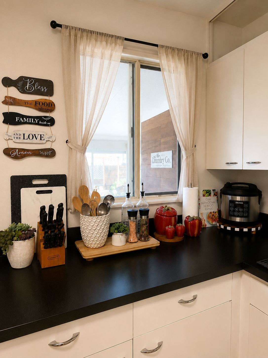 A kitchen with a window, a sign for a laundry company, a cutting board, knives, wooden spoons, salt and pepper shakers, a red pepper, and a slow cooker.