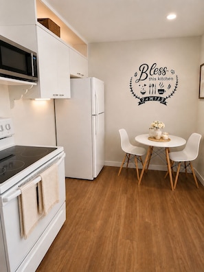Une cuisine moderne avec des appareils électroménagers blancs, une table à manger avec des chaises et un autocollant mural sur lequel on peut lire : « Que cette cuisine soit bénie, FAMILLE ».