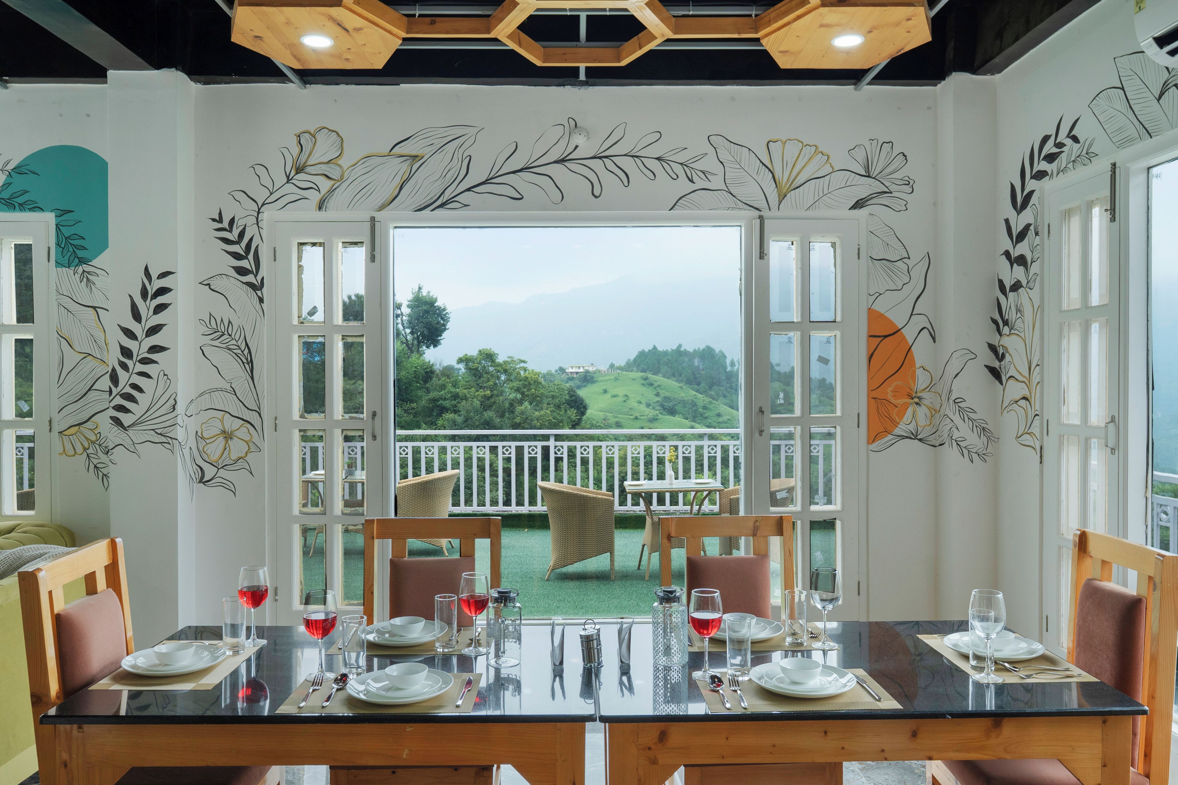 A dining area with a table set for four, a view of a green landscape through open doors, and wall art with leaf patterns.