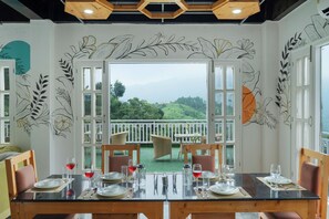 Comedor con una mesa lista para cuatro personas, vista a un paisaje verde a través de puertas abiertas y decoración mural con motivos de hojas.