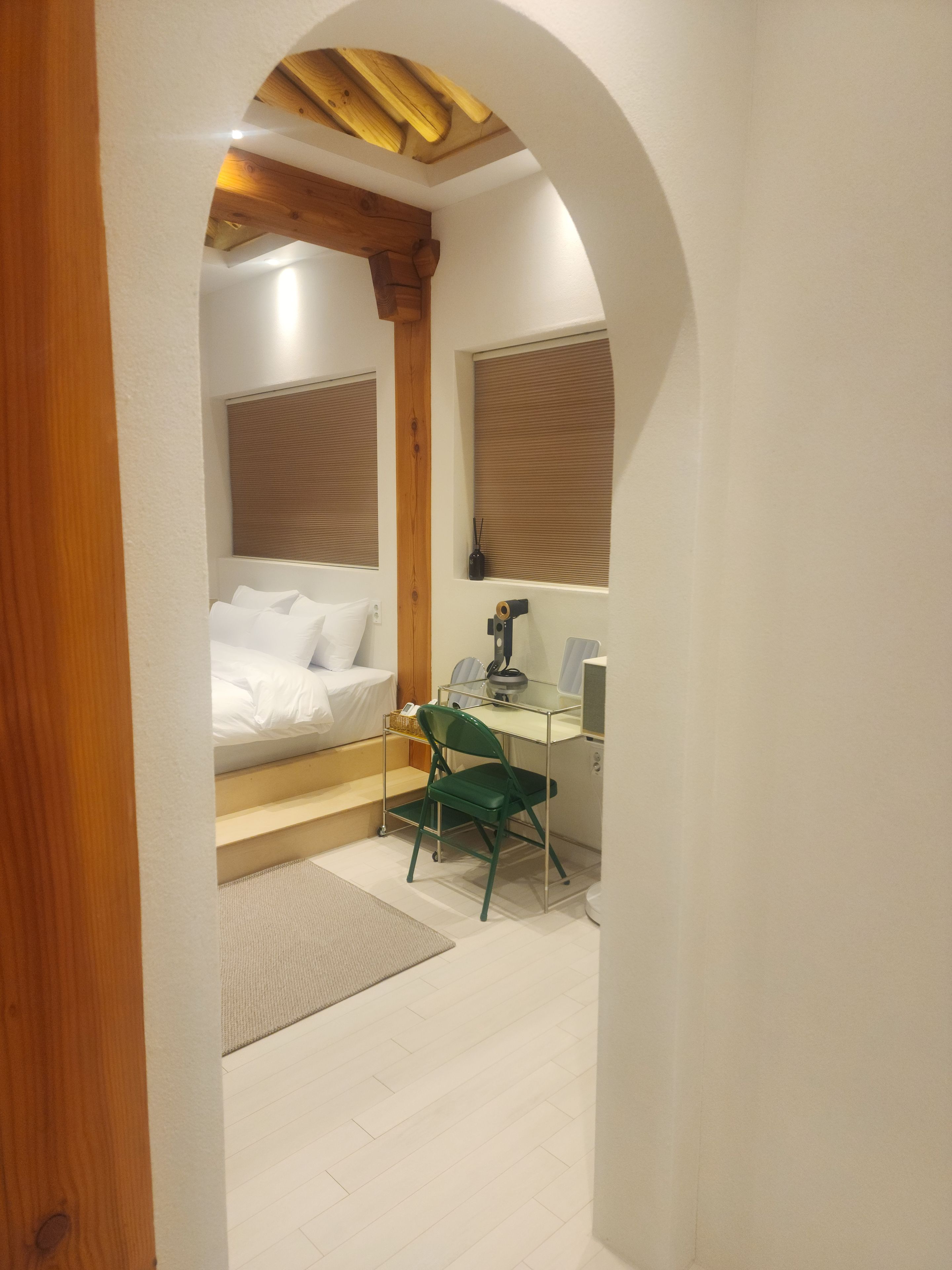 Een slaapkamer met een bed, een bureau en een stoel. Er is een raam met zonwering, een houten plafond en een vloerbedekking.