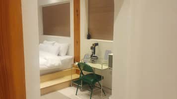 Een slaapkamer met een bed, een bureau en een stoel. Er is een raam met zonwering, een houten plafond en een vloerbedekking.