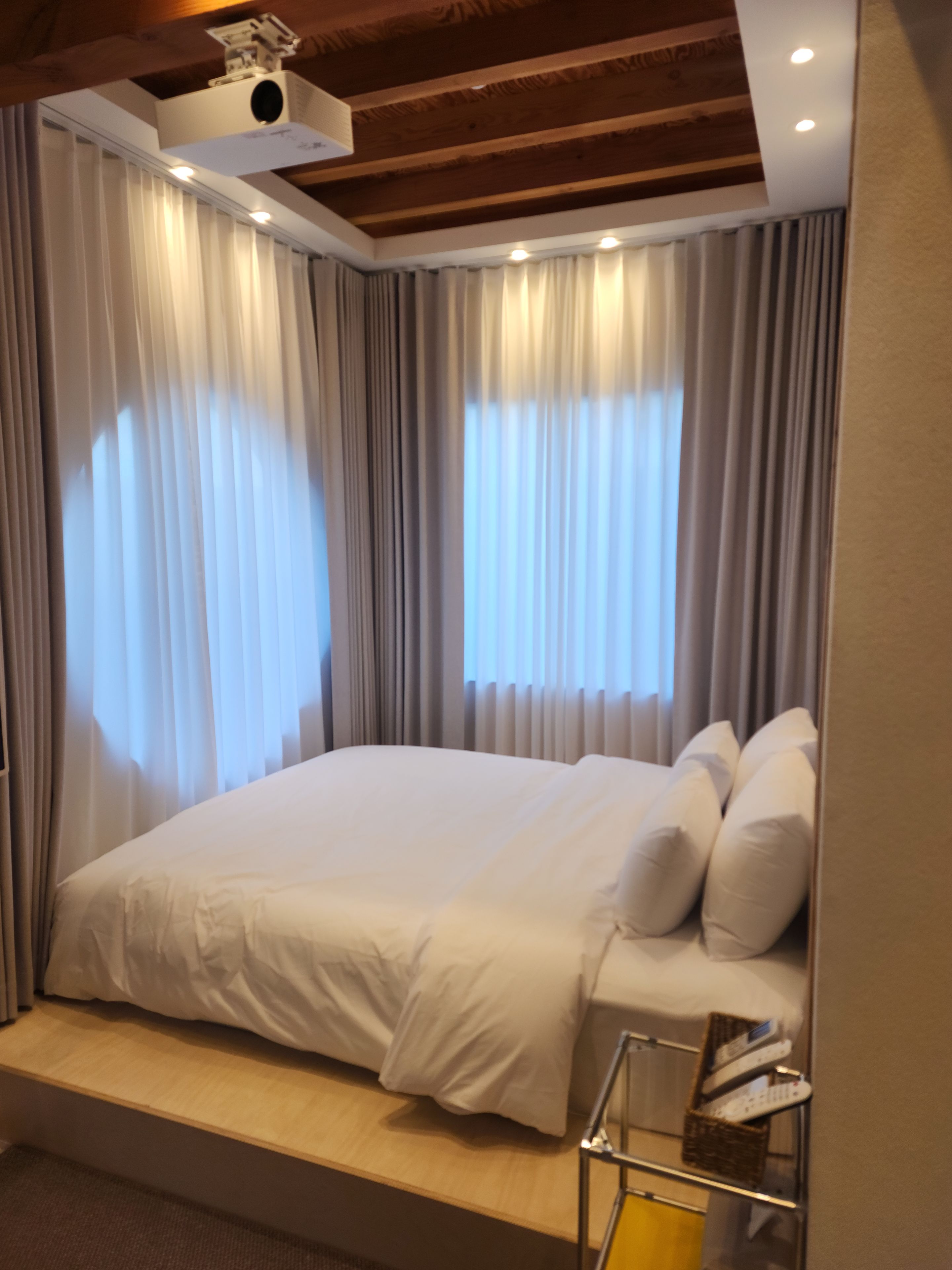 A hotel room with a bed, curtains, a ceiling projector, and a small table with a remote control.