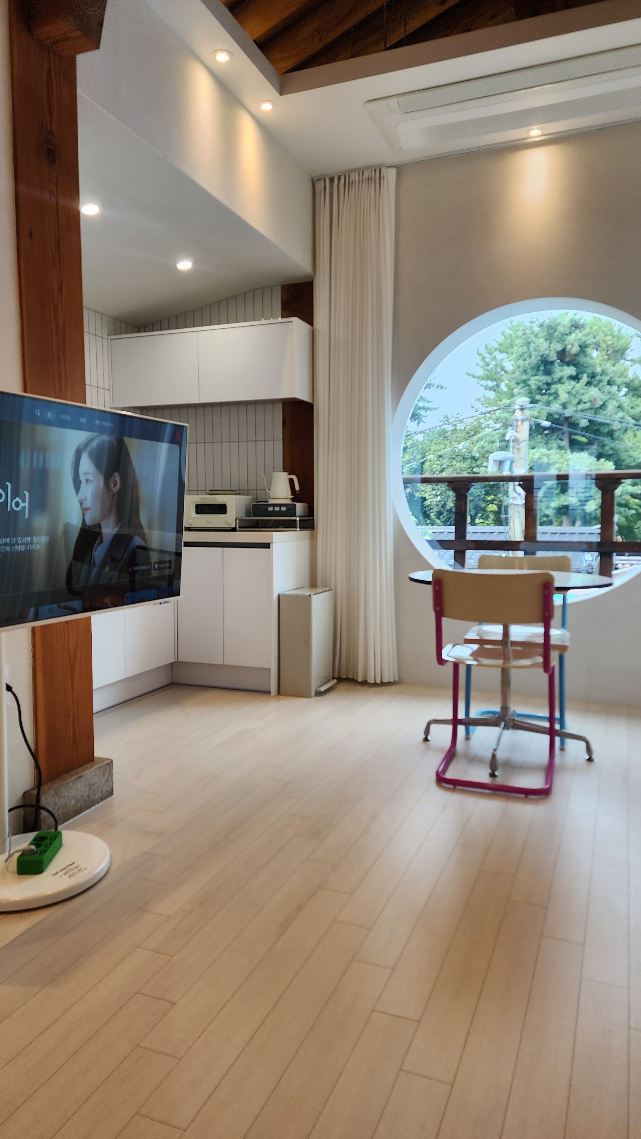A modern living room with a television, a dining area with a table and chairs, and a view of greenery through a large window.