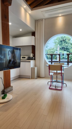 Een moderne woonkamer met een televisie, een eethoek met een tafel en stoelen, en uitzicht op groen via een groot raam.