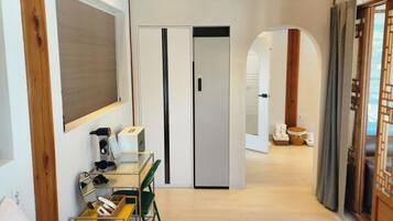 A hotel room with a bed, a desk with a chair, a mirror, and a wardrobe. There is a door leading to another room.