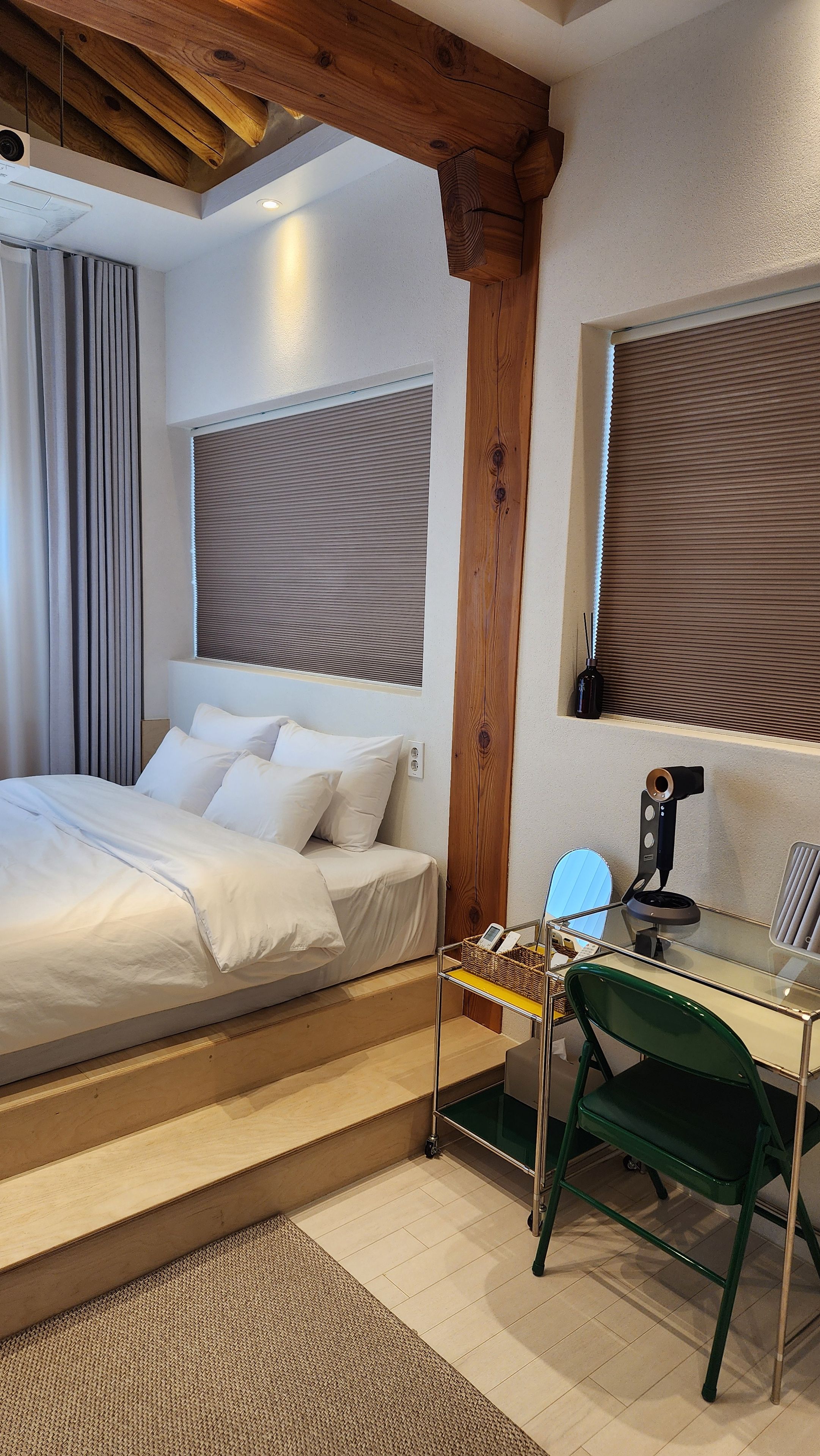 A hotel room with a bed, a desk with a chair, a window with blinds, and a wooden ceiling beam.