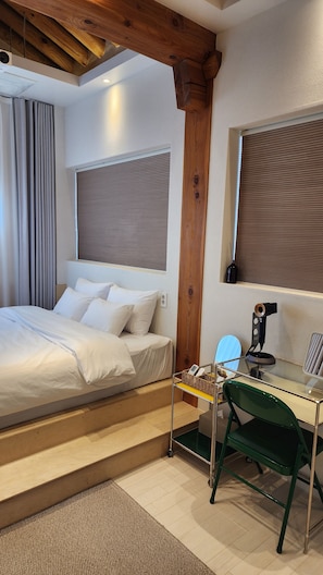 Een hotelkamer met een bed, een bureau met stoel, een raam met zonwering en een houten plafondbalk.