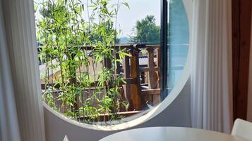 A room with a round table and chairs, a window with a view of greenery and a building outside.