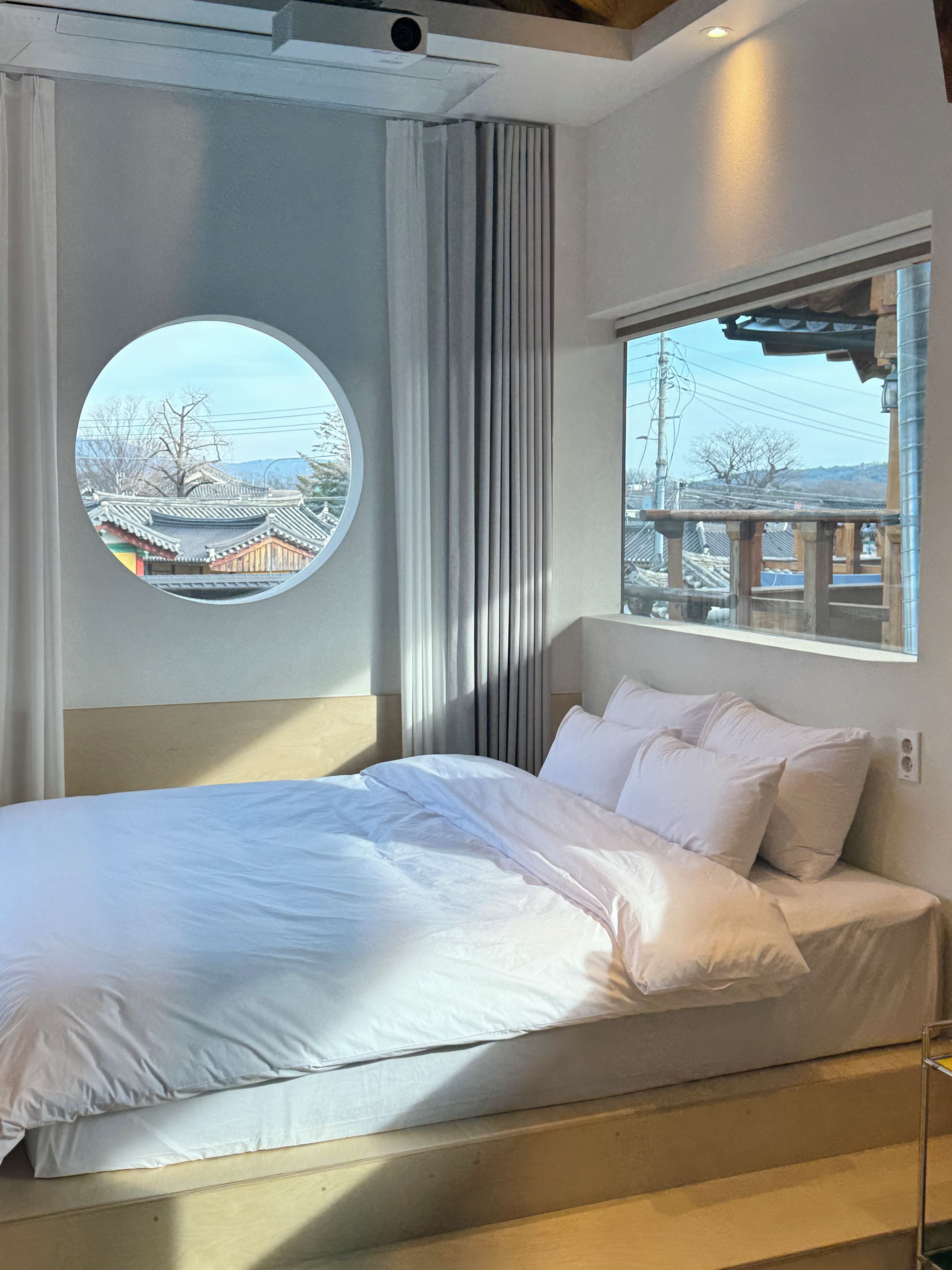 A hotel room with a bed, a round window with a view of traditional buildings, and a window with a view of a bridge.