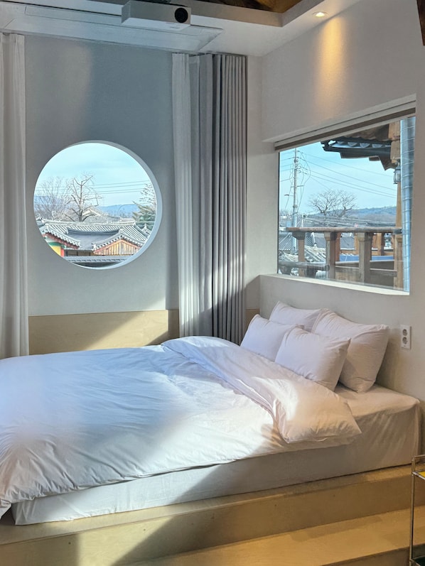 A hotel room with a bed, a round window with a view of traditional buildings, and a window with a view of a bridge.