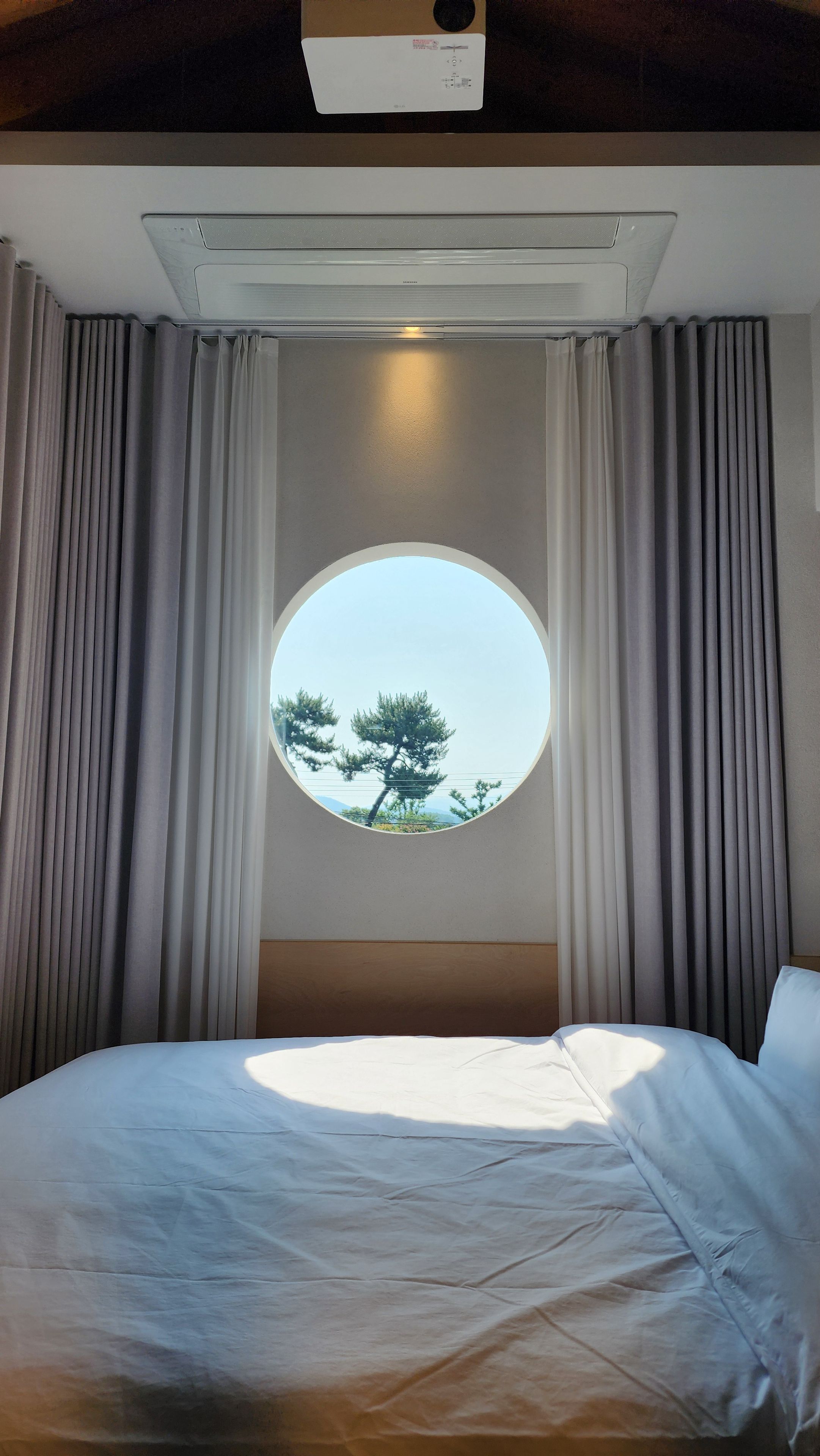 A hotel room with a round window, curtains, a bed, and an air conditioning unit.