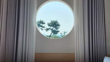 A hotel room with a round window, curtains, a bed, and an air conditioning unit.