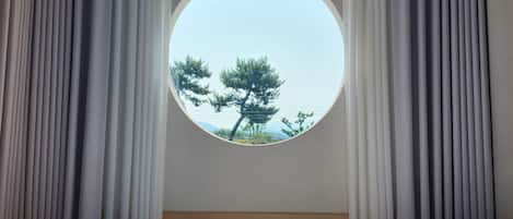 A hotel room with a round window, curtains, a bed, and an air conditioning unit.