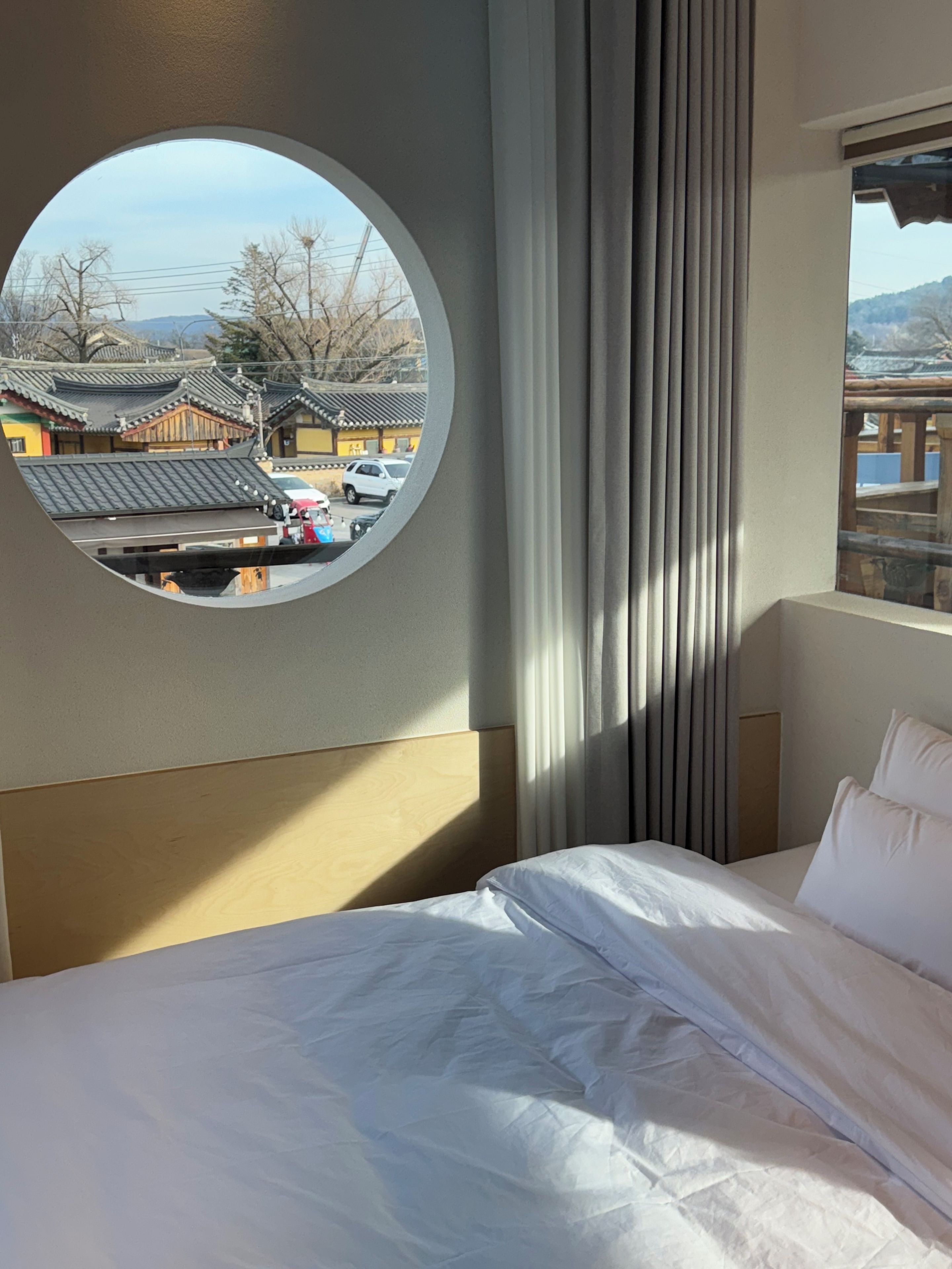A hotel room with a bed, a round window with a view of traditional buildings, and a curtain.