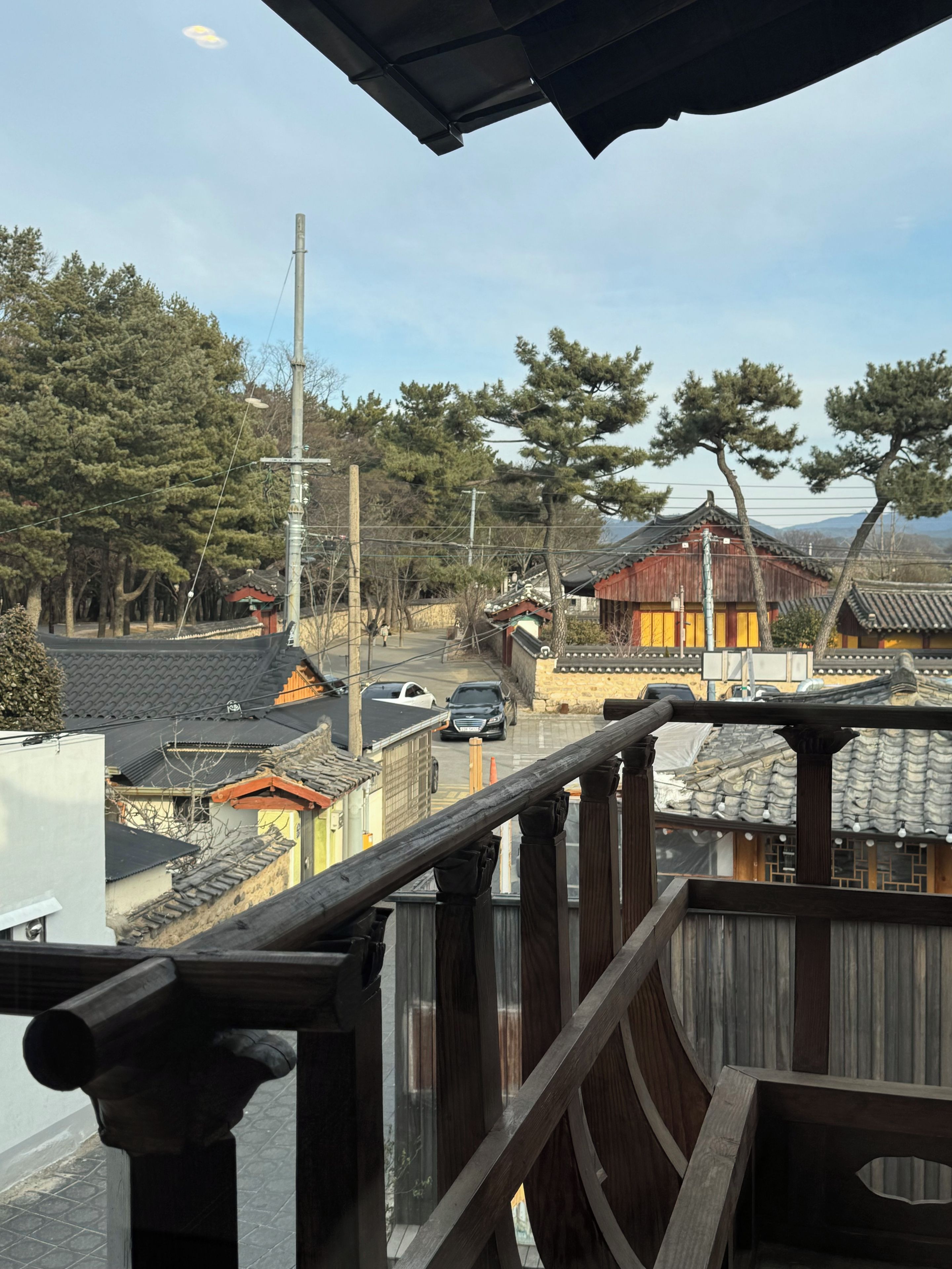 A traditional wooden building with a tiled roof, surrounded by trees and a street with parked cars.