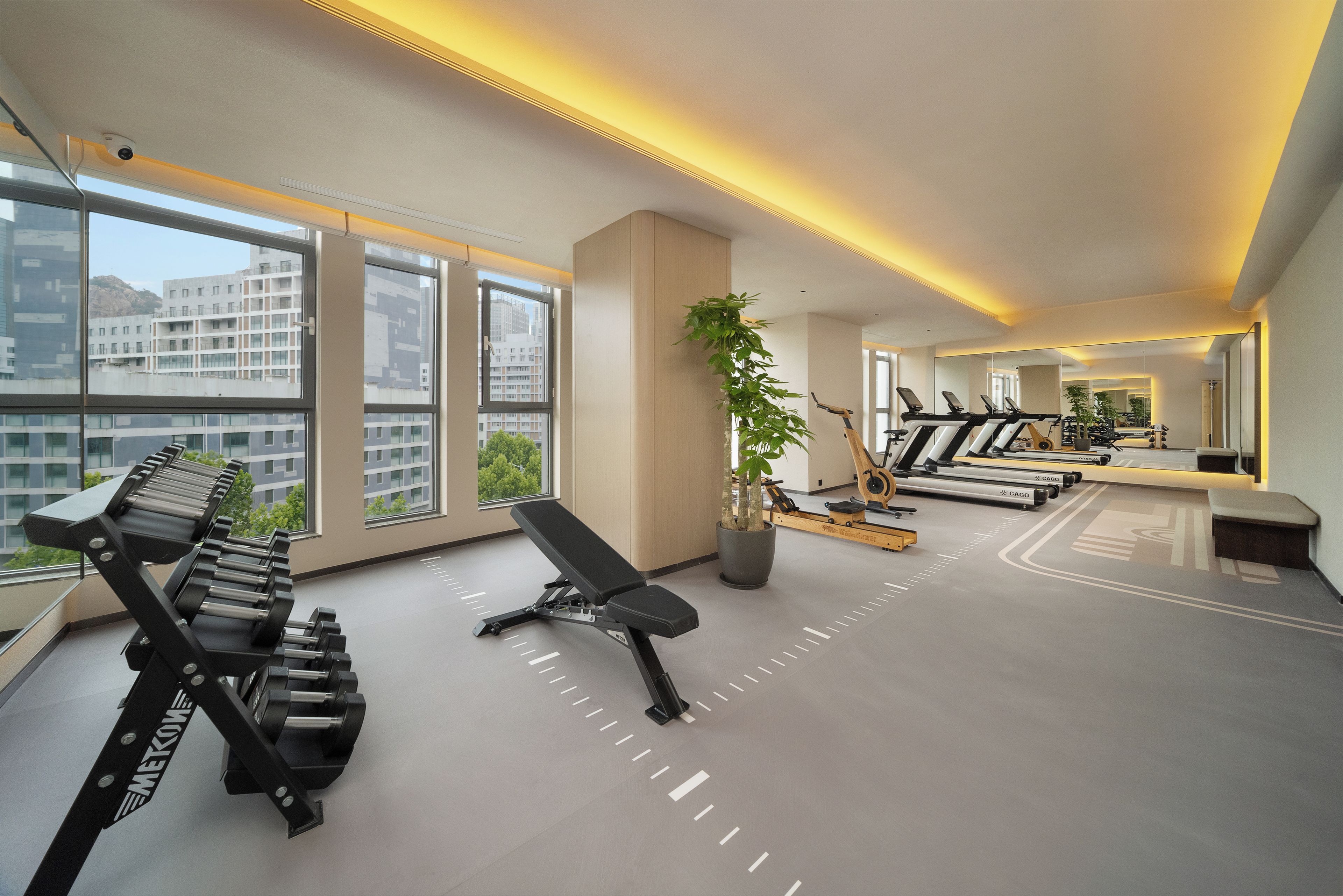 A modern gym with exercise equipment, large windows, and a view of buildings.