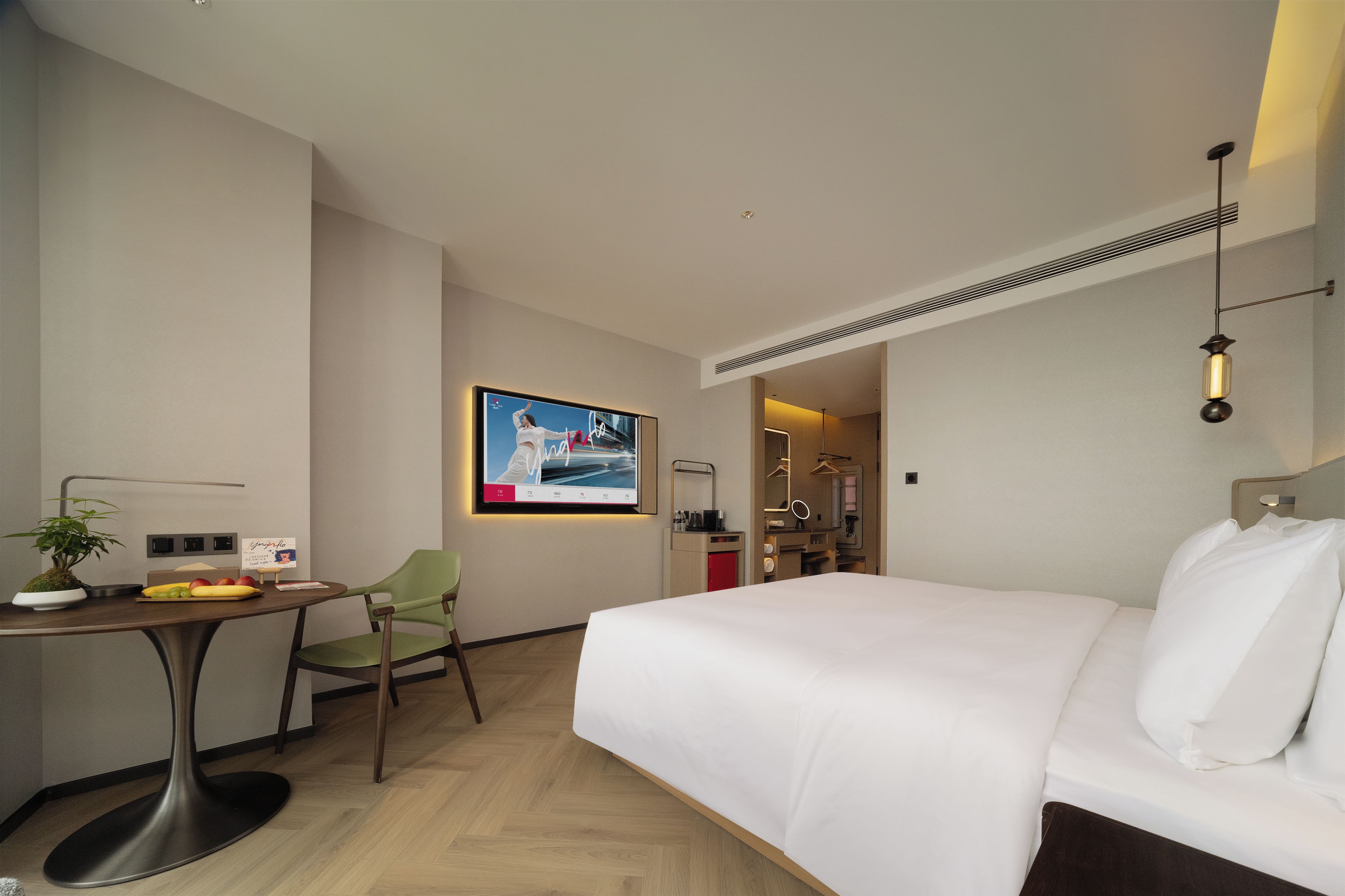 A hotel room with a bed, a desk, a chair, a small table with a plant, and a wall-mounted TV.
