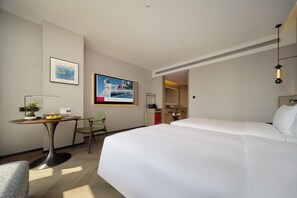 A hotel room with a bed, a desk with a chair, a small table with fruits, a potted plant, and a painting on the wall.