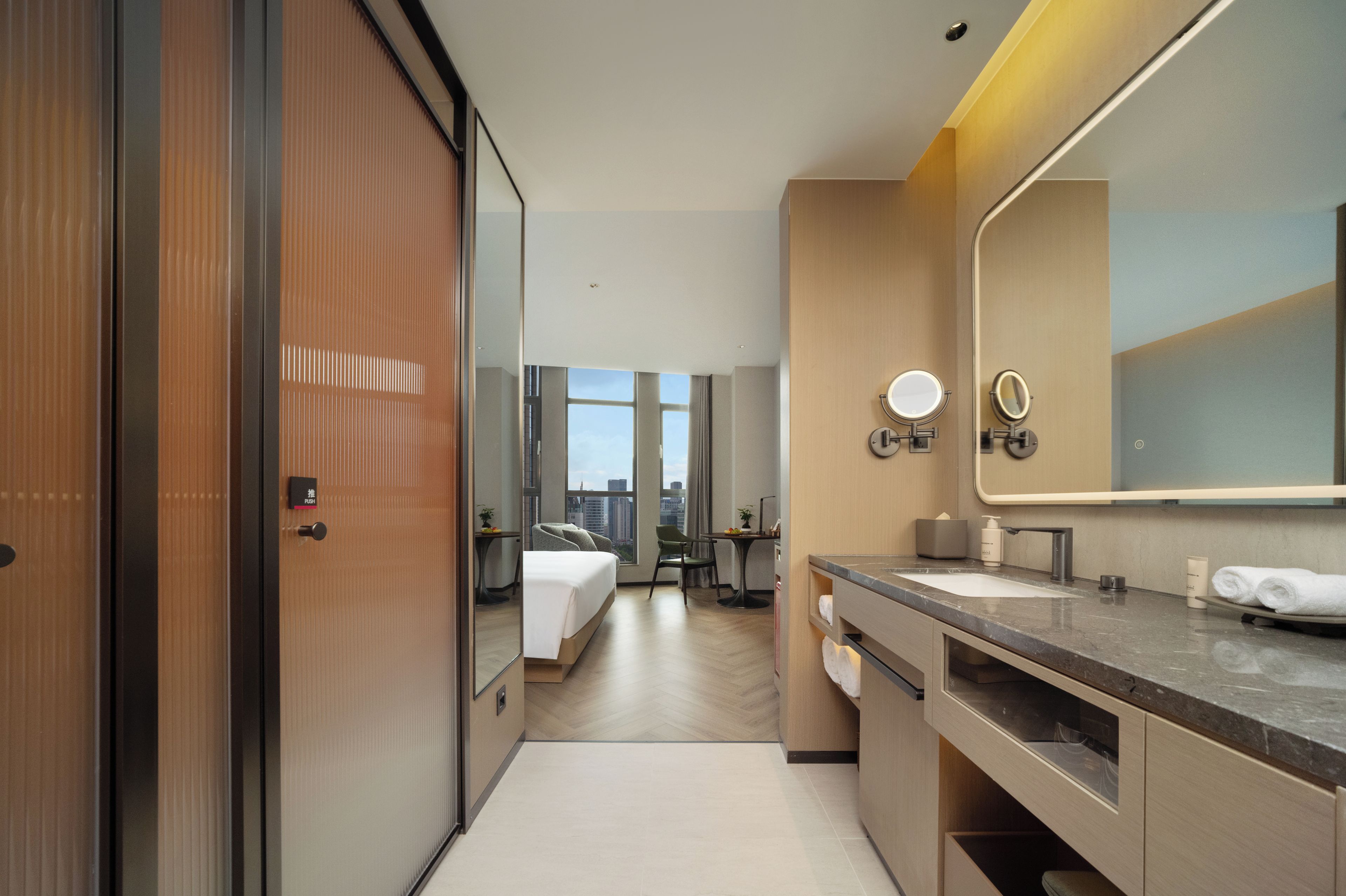 A hotel room with a bed, a bathroom with a mirror and sink, and a view of the city through large windows.