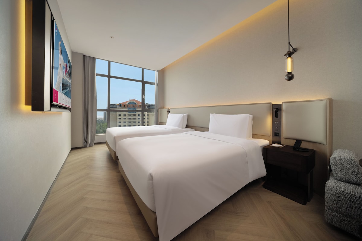 A hotel room with two beds, a large window, a desk with a lamp, and a chair.
