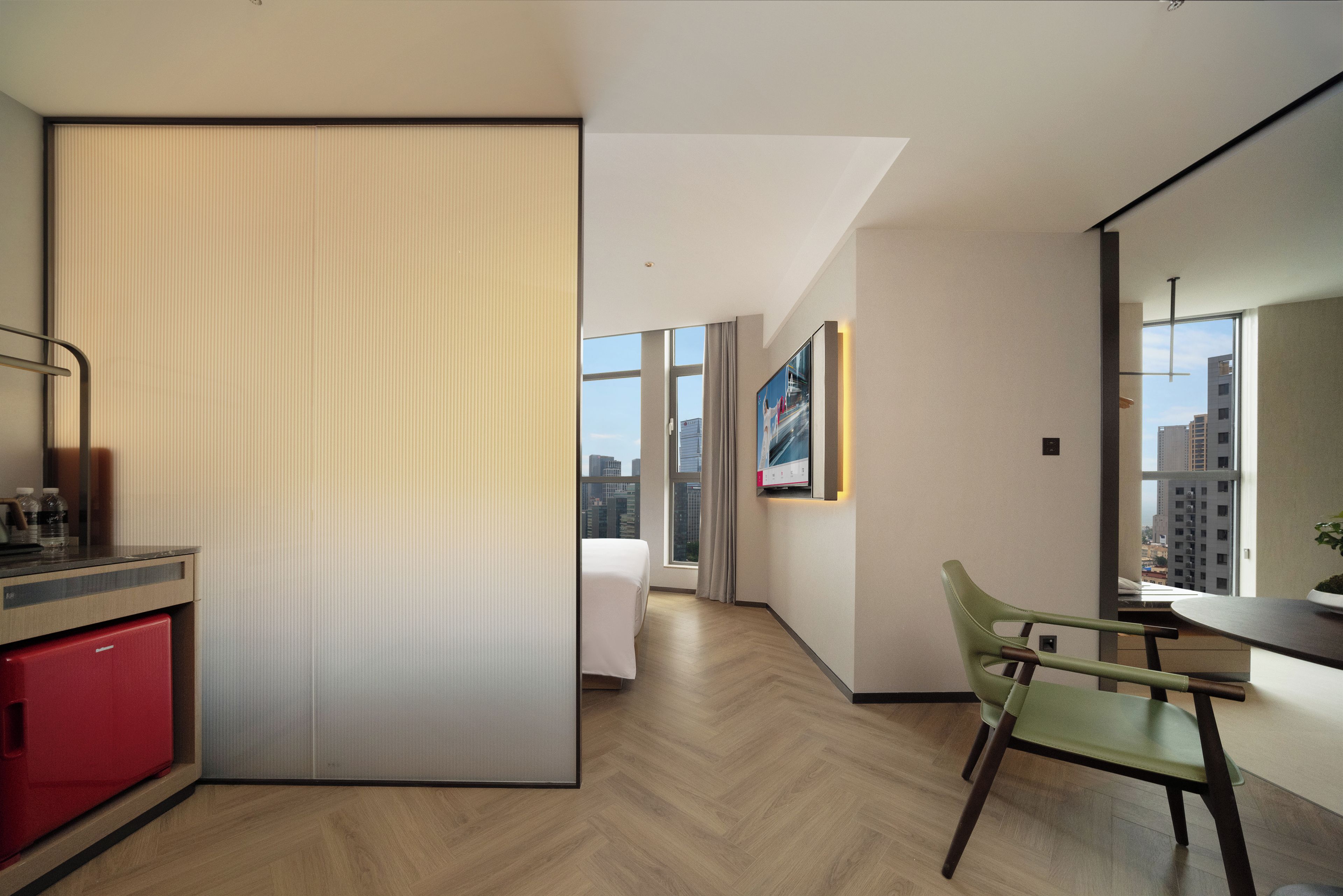 A modern hotel room with a sliding door, a dining area with a green chair, and a view of the cityscape through large windows.