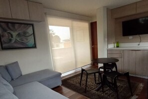 Ett vardagsrum med en soffa, ett matbord med stolar, en platt-TV som är monterad på väggen och en skjutdörr i glas som leder ut till ett utomhusområde.
