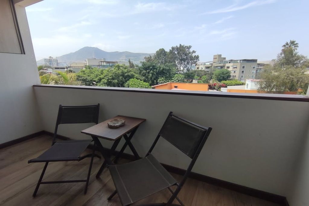 Un balcón con dos sillas y una mesa pequeña, con vista a un paisaje urbano con edificios y árboles.