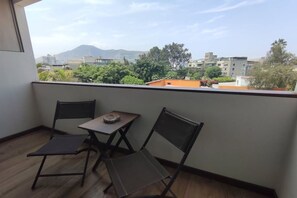 Un balcón con dos sillas y una mesa pequeña, con vista a un paisaje urbano con edificios y árboles.
