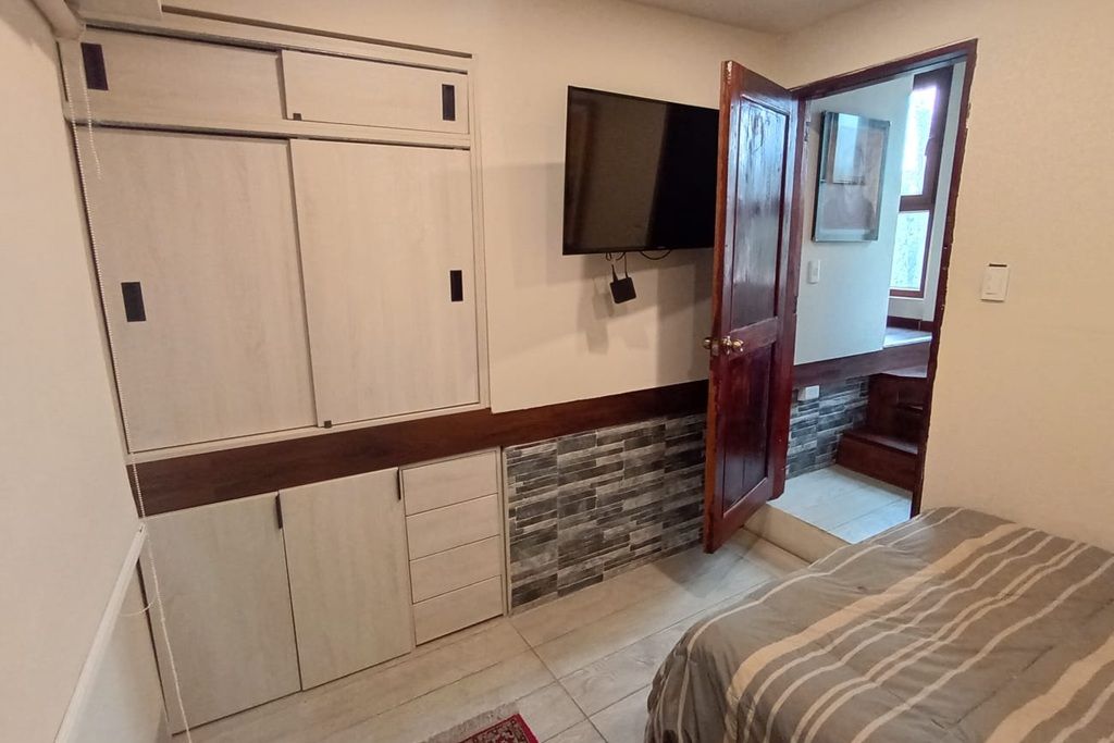 Ein Schlafzimmer mit Bett, Kleiderschrank, Fernseher und einem durchgehenden Türrahmen sichtbaren Badezimmer.