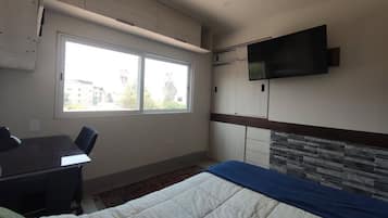 A bedroom with a bed, a desk, a chair, a television, and a window.