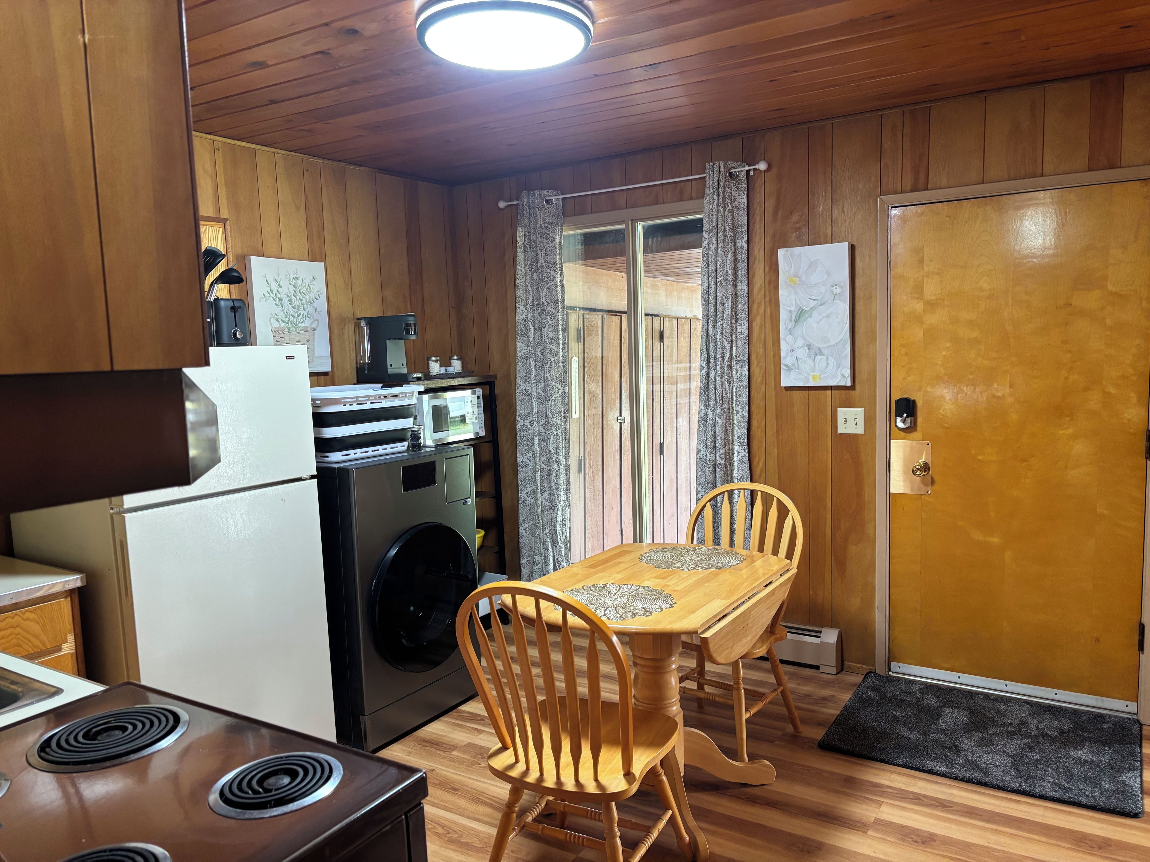 A compact kitchen with a refrigerator, microwave, and washing machine. There is a wooden dining table with chairs and a door with a doormat.