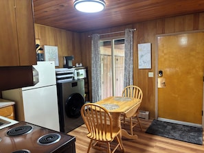 A compact kitchen with a refrigerator, microwave, and washing machine. There is a wooden dining table with chairs and a door with a doormat. - Cozy, Affordable Retreat Near Anchorage Airport (Anchorage)