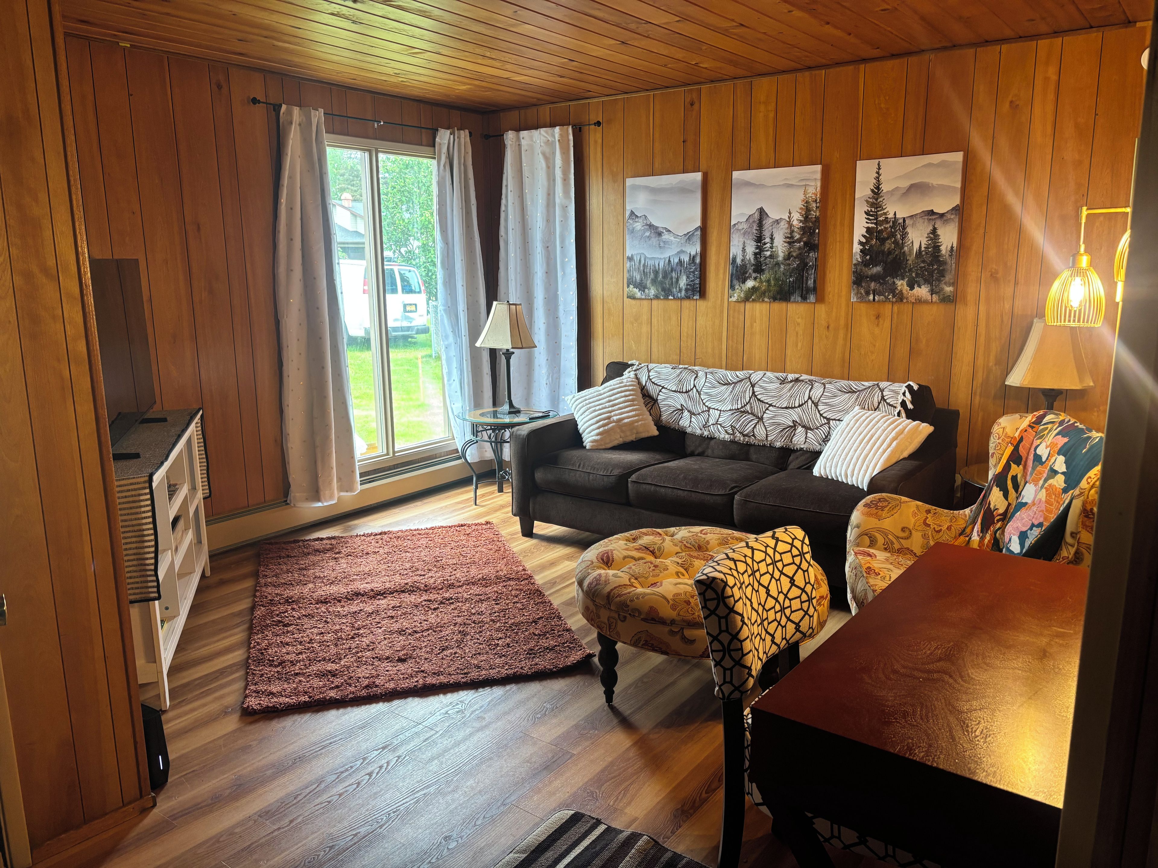A cozy living room with a sofa, armchair, and a coffee table. There is a window with curtains, a lamp, and artwork on the wall.