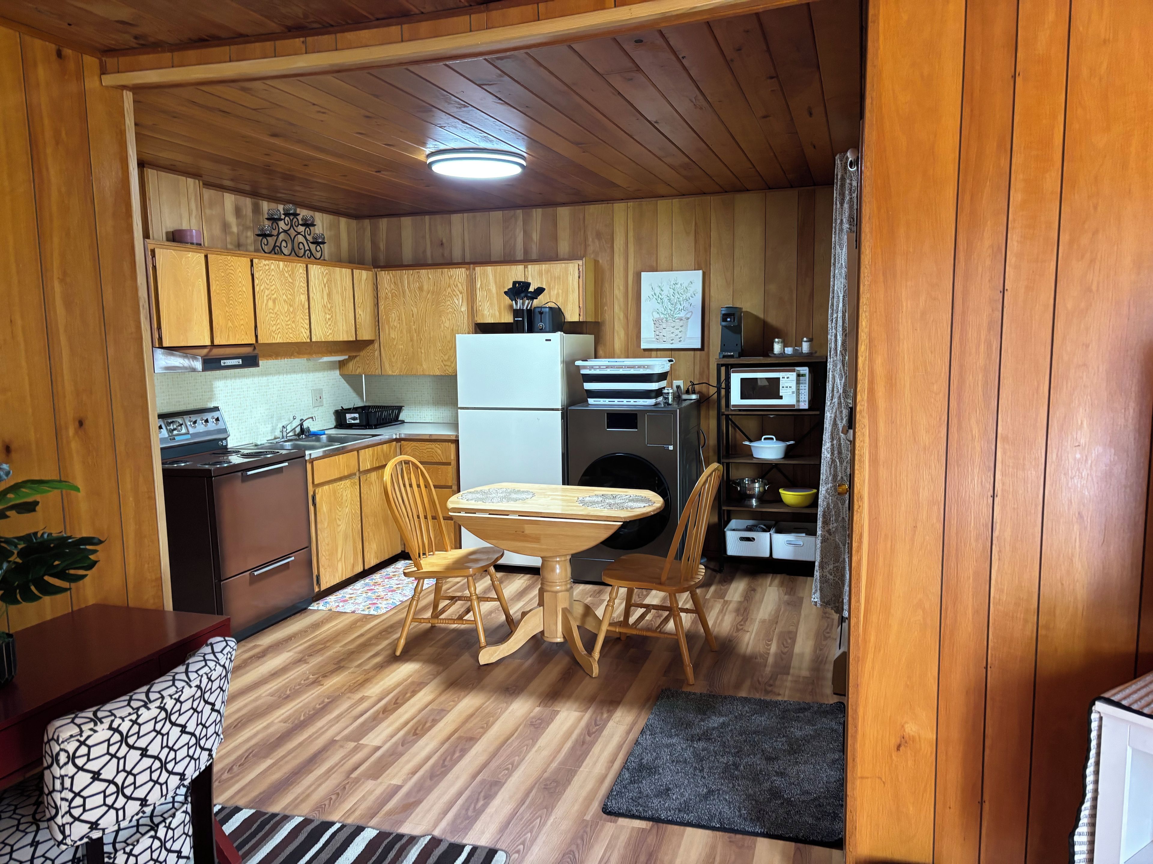 A compact kitchen with wooden cabinets, a dining table with chairs, a refrigerator, a microwave, and a washing machine.